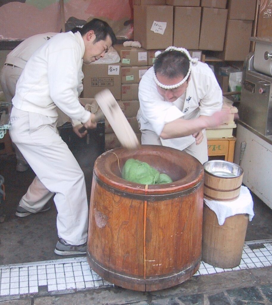 Two men in white overalls take it in rapid turn to hammer something green inside a large wooden mortar.
