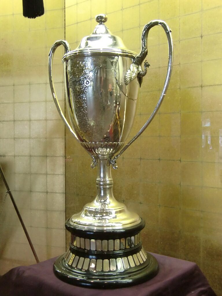 A colour photo of a large metal trophy, with large handles. It stands on a black base with many small metal tokens on it. The trophy is intricately engraved, but the details aren't clear from the angle of the photograph.