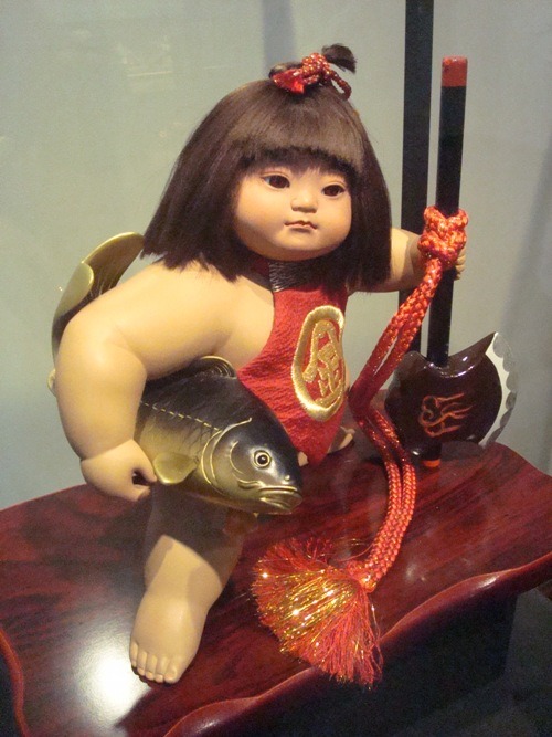 A colour image of a statue. A short Japanese child, carrying a fish under one arm, and holding an axe with the other, looks into the distance. He has brown hair tied in the samurai topknot, and he wears an apron with the Japanese character for "gold" written on it, surrounded by a circle.