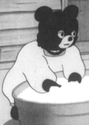 A dopey-eyed bear, wearing a jumper, reaches into a large bowl. They're washing something, either clothes or plates.