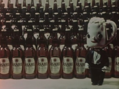A colour stop-motion image. A human figure with the head of a beer stein with human features stands in front of a huge array of beer bottles.